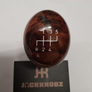 Può includere: Un pomello del cambio in legno con finitura marrone scuro venata. Lo schema del cambio è bianco, con i numeri 1, 2, 3, 4, 5 e R. Il logo "JACKKNOBZ" è visibile su una carta nera sottostante.