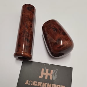 Puede incluir: Dos pomos de palanca de cambios de madera con acabado brillante. Uno es cilíndrico, el otro tiene forma ovalada. Ambos son de un color marrón rojizo intenso con un patrón de vetas de madera único. Una tarjeta de visita con el texto "JACKKNOBZ" está en primer plano.
