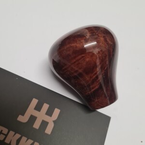 Può includere: Un pomello del cambio lucido, a forma di goccia, con un ricco motivo a venature di legno marrone rossiccio. Il pomello è liscio e lucido, riflettendo la luce. Una carta scura con il testo "CKKNOBZ" e un logo è parzialmente visibile.