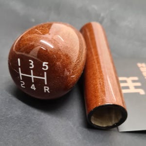 May include: A wooden gear stick knob and a matching cylindrical handle. The knob is brown with white numbers and a gear pattern. The handle is a similar shade of brown. Automotive parts.