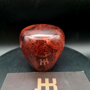Peut inclure: Un pommeau de levier de vitesse en forme de goutte, poli, fabriqué en bois brun rougeâtre avec un motif de grain sombre et tourbillonnant. La surface supérieure présente une grille de vitesse gravée et la marque JOCKKNOBZ est imprimée en dessous.