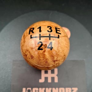 May include: A spherical wooden gear shift knob with a polished finish and a natural brown wood grain pattern. The top surface features a black printed gear shift pattern with the letters R, 1, 3, E and the numbers 2, 4 arranged in a grid.