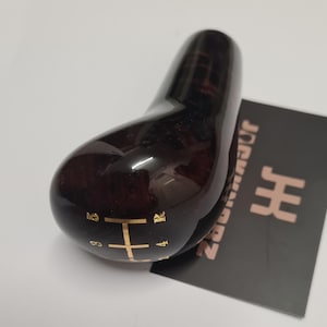Könnte beinhalten: Ein dunkelbrauner, glänzender Schalthebelknauf mit goldfarbenen Gangmarkierungen. Der Knauf hat eine glatte, ergonomische Form und liegt auf einer schwarzen Karte mit goldenem Text und einem Logo.