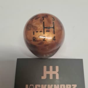 Può includere: Un pomello del cambio sferico marrone con un motivo a venature del legno. Lo schema del cambio è stampato in nero, con i numeri da 1 a 4 e R per la retromarcia. Il logo "JACKKNOBZ" è stampato su una scheda nera.