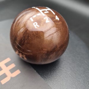 Könnte beinhalten: Ein kugelförmiger Schaltknauf mit polierter brauner Holzmaserung. Die Oberseite zeigt weiße aufgedruckte Schaltschema-Markierungen, einschließlich der Buchstaben R, 2 und 4, sowie gestrichelte Linien, die den Schaltweg für ein Schaltgetriebe angeben.