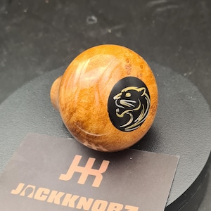 May include: A spherical wooden gear shift knob with a polished finish and a prominent wood grain pattern. A circular black inlay on the side features a stylized gold-colored panther head graphic. A card below displays the text JACKKNORZ in gold.