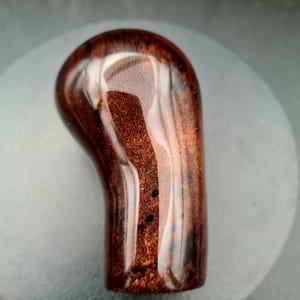 May include: A polished, dark brown wooden gear shift knob with a smooth, glossy finish. The wood features a rich, swirling grain pattern with deep mahogany tones and subtle black speckling throughout the curved, ergonomic, cylindrical shape.