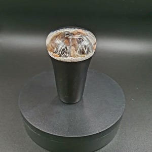 May include: A black and clear resin gear shift knob with a unique design. The knob is on a black circular base.