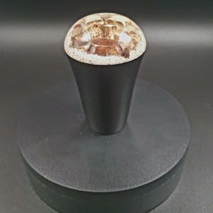 May include: A black wooden knob with a clear resin top containing a skull design. The knob is on a black circular base.