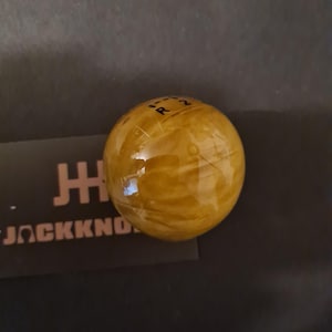 Può includere: Un pomello del cambio sferico giallo dorato con finitura lucida. La parte superiore del pomello presenta segni neri che indicano le posizioni delle marce. Il logo "JACKKNO" è visibile su uno sfondo scuro.