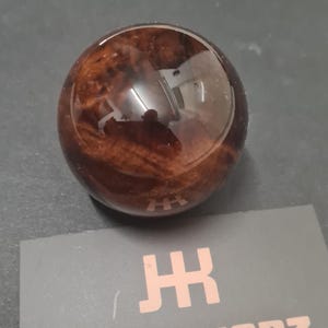 Puede incluir: Un pomo de cambio redondo de madera marrón con un acabado brillante. La veta de la madera es visible y el pomo se encuentra sobre una superficie gris. Una tarjeta de visita con el texto "JACKKNOBZ" y un logotipo está en primer plano.
