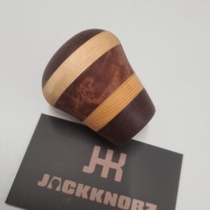 Könnte beinhalten: Ein hölzerner Schalthebelknauf mit abgerundeter Oberseite und zylindrischer Basis. Der Knauf hat abwechselnde Bänder aus dunkelbraunem und hellbraunem Holz. Eine Visitenkarte mit dem Text "JACKKNOBZ" befindet sich im Vordergrund.