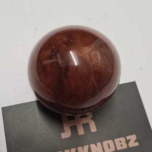 May include: A glossy, spherical, brown shift knob with a wood grain pattern. The shift knob is sitting on a black card with the text "JACKKNOBZ" in copper-colored letters.