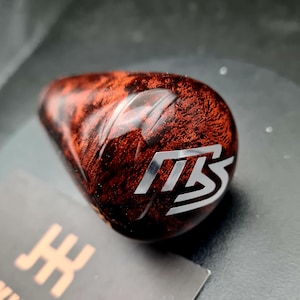 Può includere: Un pomello del cambio a forma di goccia con una venatura del legno radica lucidata in un intenso color marrone rossastro e una finitura lucida. La superficie superiore presenta un logo metallico color argento con le lettere TS stilizzate.