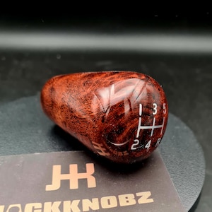 May include: A polished, reddish-brown burl wood gear shift knob with a smooth, glossy finish. The top surface features a white engraved manual transmission shift pattern showing gears 1, 3, 5, 2, 4, and R. The logo JK is etched on the side.