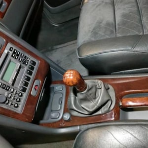 May include: The interior of a vehicle featuring a wooden gear shift knob with a manual transmission pattern, a grey leather gear shift boot, and a wooden center console trim. The dashboard includes a radio unit with buttons and a digital display.