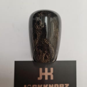 May include: A glossy, dark brown shift knob with a swirling, wood-grain pattern. The knob sits atop a black card with the text "JACKKNOBZ" and a stylized logo in a copper color.