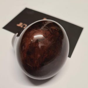 Könnte beinhalten: Ein glänzendes, dunkelbraunes, oval geformtes Objekt mit Holzmaserung. Das Objekt hat eine glatte, reflektierende Oberfläche und scheint ein dekoratives Element oder Teil eines größeren Produkts zu sein. Der Hintergrund ist eine weiße Oberfläche mit einer schwarzen Karte.