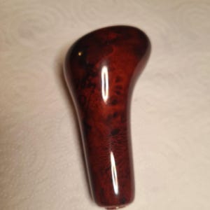 May include: A polished gear shift knob featuring a deep reddish-brown wood grain pattern with dark swirling details. The object has a smooth, glossy finish and an asymmetrical, ergonomic shape that tapers toward the base.