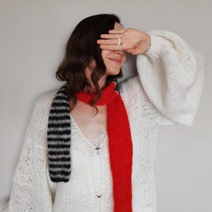 Puede incluir: Un suéter de punto blanco grueso con mangas largas abullonadas y cuello en V, combinado con una bufanda larga que presenta un lado rojo sólido y un lado con rayas horizontales negras y grises, usado sobre una camiseta blanca con encaje.