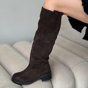 May include: Brown suede knee-high boots with a chunky heel and a buckle detail on the side.