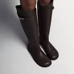 May include: A pair of dark brown knee-high boots featuring a smooth finish, a rounded toe, and a thin strap detail encircling the upper shaft. The boots are secured with a small metallic buckle on the outer side and have a flat, dark sole.