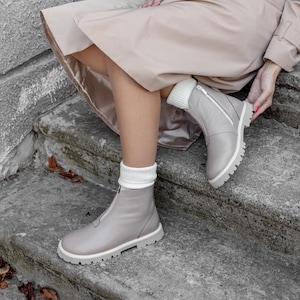 May include: Light gray leather boots with white soles and a zipper closure. The boots have a chunky, lugged sole and are paired with white knit socks. The boots are part of a fall fashion ensemble.