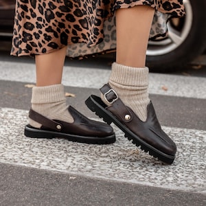 Puede incluir: Un par de sandalias de cuero marrón con una correa de hebilla y una suela gruesa. Las sandalias se llevan con calcetines beige y una falda de estampado de leopardo.