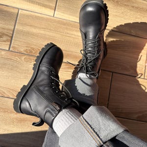 Könnte beinhalten: Schwarze Leder-Combat-Stiefel mit Schnürung und seitlichem Reißverschluss. Die Stiefel haben eine dicke Profilsohle und werden mit grauen Jeans und weißen Socken getragen. Die Stiefel stehen auf einem Holzboden.