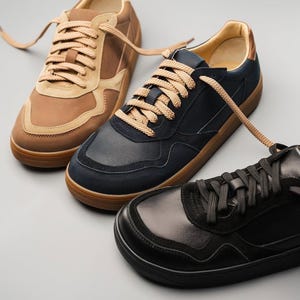 May include: Three pairs of sneakers in brown, navy, and black. The brown pair has tan suede accents and light brown laces. The navy pair has a suede toe and light brown laces. The black pair has black laces and a smooth leather finish.