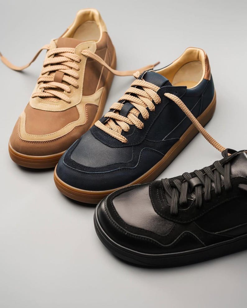 May include: Three pairs of sneakers in brown, navy, and black. The brown pair has tan suede accents and light brown laces. The navy pair has a suede toe and light brown laces. The black pair has black laces and a smooth leather finish.