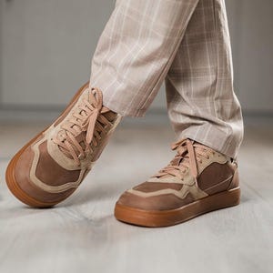 May include: Brown and tan sneakers with matching laces and a brown sole. The sneakers are worn with tan plaid pants. The shoes have a layered design with a mix of textures and shades of brown.