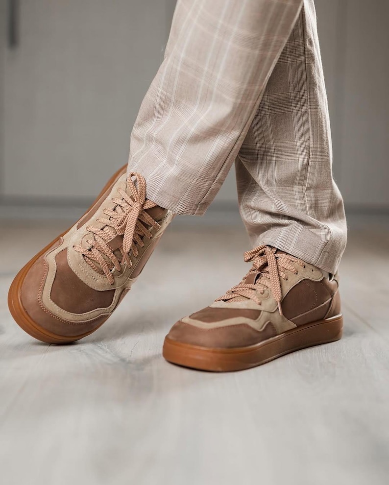 May include: Brown and tan sneakers with matching laces and a brown sole. The sneakers are worn with tan plaid pants. The shoes have a layered design with a mix of textures and shades of brown.
