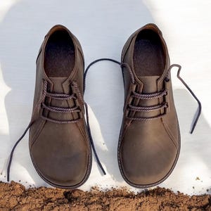 May include: A pair of brown leather shoes with laces, presented on a white surface with a small pile of dirt at the bottom. The shoes have a classic design with a rounded toe and a textured finish.