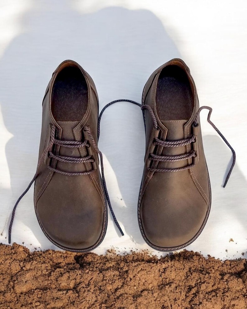 May include: A pair of brown leather shoes with laces, presented on a white surface with a small pile of dirt at the bottom. The shoes have a classic design with a rounded toe and a textured finish.