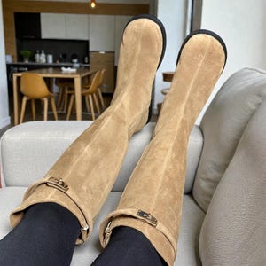 May include: A pair of knee-high tan suede boots featuring a rounded toe, a black sole, and a decorative strap across the upper shaft secured with a gold-toned metal buckle hardware. The boots are worn over black leggings, extending to the mid-calf.