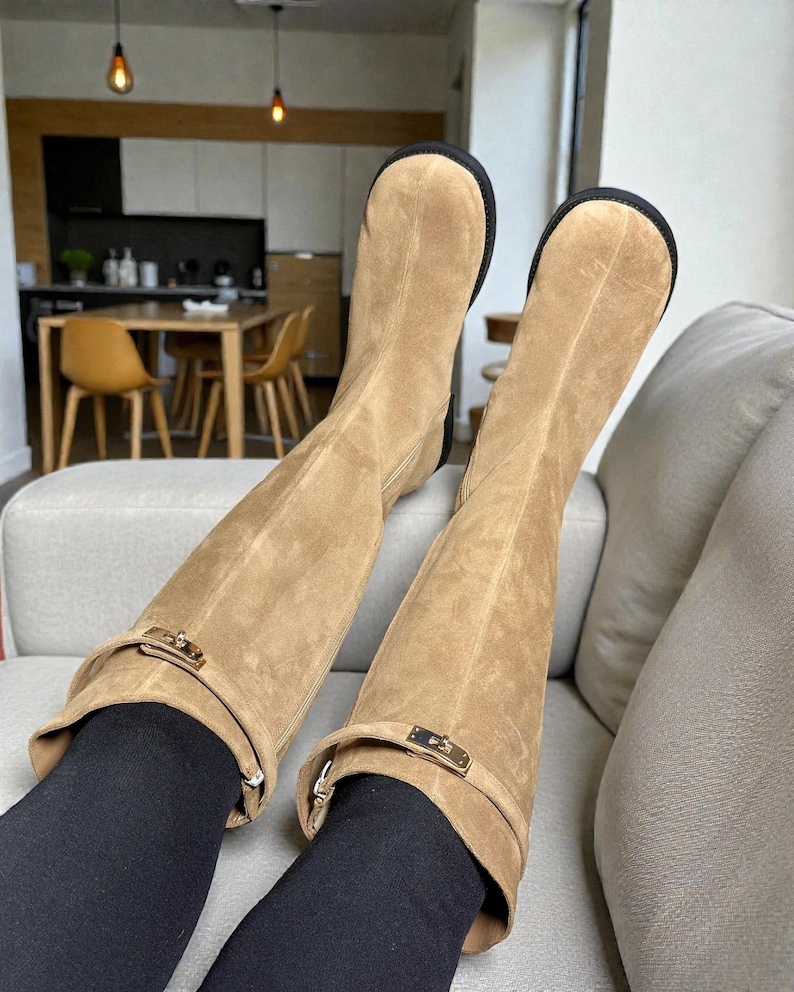 May include: A pair of knee-high tan suede boots featuring a rounded toe, a black sole, and a decorative strap across the upper shaft secured with a gold-toned metal buckle hardware. The boots are worn over black leggings, extending to the mid-calf.