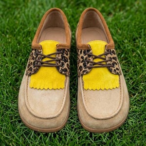 May include: A pair of slip-on shoes featuring a tan suede base, a bright yellow fringed tongue, and a leopard print panel around the lace-up section. The shoes include brown laces, metal eyelets, and a brown suede trim along the collar opening.