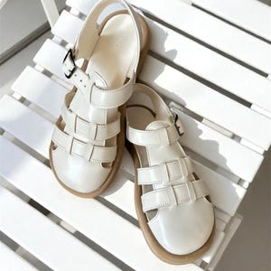 May include: A pair of off-white leather sandals with a cage-like design. The sandals have a buckle closure and a light brown sole. The shoes are sitting on a white chair.