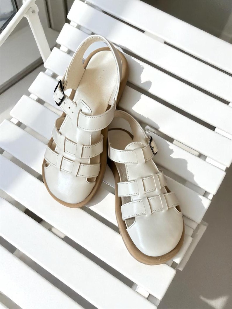 May include: A pair of off-white leather sandals with a cage-like design. The sandals have a buckle closure and a light brown sole. The shoes are sitting on a white chair.