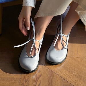 Op de afbeelding: Lichtblauwe Mary Jane-schoenen met een ronde neus en zwarte zolen. De schoenen hebben een bandje dat om de enkel wordt gestrikt. De schoenen zijn gemaakt van glad materiaal.