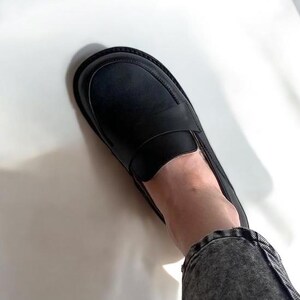 May include: Black leather loafer with a rounded toe and a thick sole. The shoe features a strap across the top and is worn with denim jeans. The shoe is a classic style.