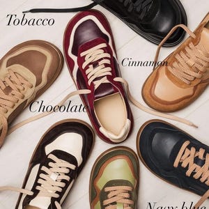May include: A collection of seven pairs of sneakers in various colors, including black, burgundy, tobacco, chocolate, cinnamon, avocado, and navy blue. Each pair features matching laces and a neutral-colored interior.