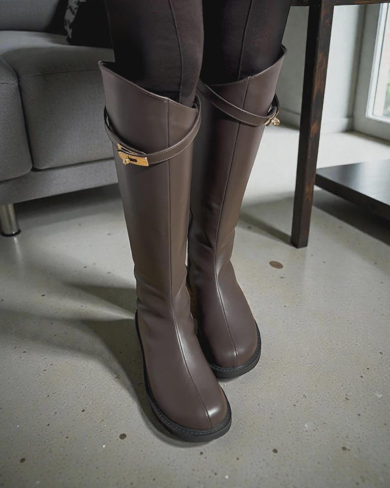 May include: A pair of knee-high brown boots featuring a smooth finish, a rounded toe, and a decorative strap across the upper shaft secured with a gold-toned metal buckle hardware. The boots have a thick, dark sole and a vertical seam down the front.