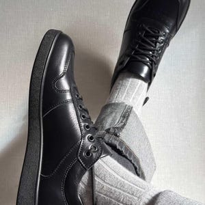 May include: A pair of black leather sneakers with laces. The shoes have a rounded toe and a thick, black sole. The image also shows gray jeans and striped socks.
