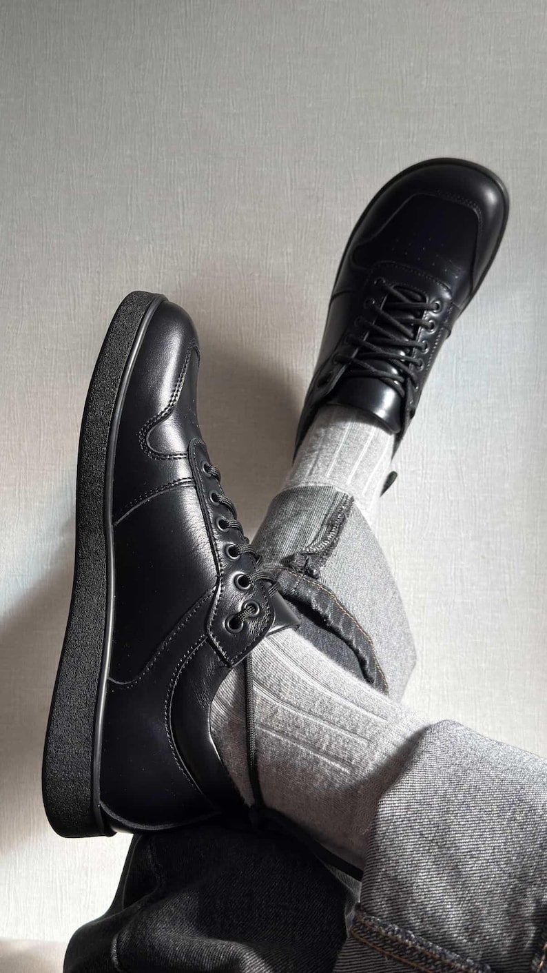 May include: A pair of black leather sneakers with laces. The shoes have a rounded toe and a thick, black sole. The image also shows gray jeans and striped socks.