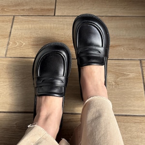 Puede incluir: Un par de mocasines negros de cuero con punta redondeada, una tira cl&aacute;sica con ranura sobre el empeine y una suela gruesa de goma texturizada. Los zapatos tienen un acabado liso y se llevan con pantalones vaqueros claros de bajo deshilachado.