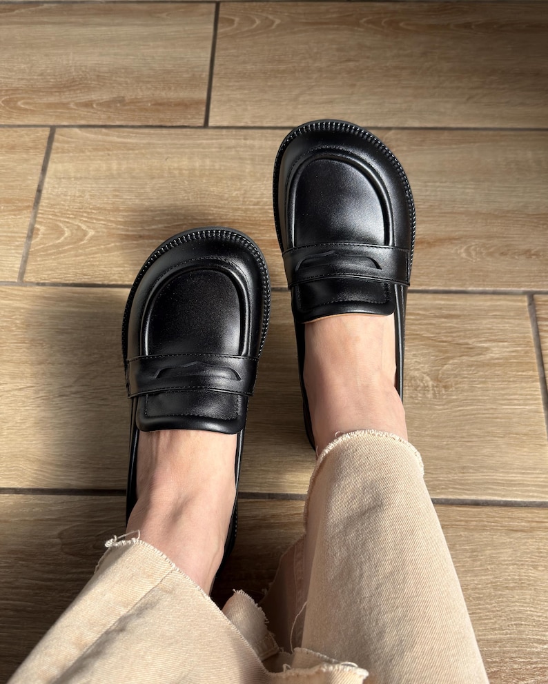 Puede incluir: Un par de mocasines negros de cuero con punta redondeada, una tira cl&aacute;sica con ranura sobre el empeine y una suela gruesa de goma texturizada. Los zapatos tienen un acabado liso y se llevan con pantalones vaqueros claros de bajo deshilachado.