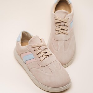 May include: A pair of light pink suede sneakers with blue and pink stripes on the sides. The laces are beige and the soles are white.