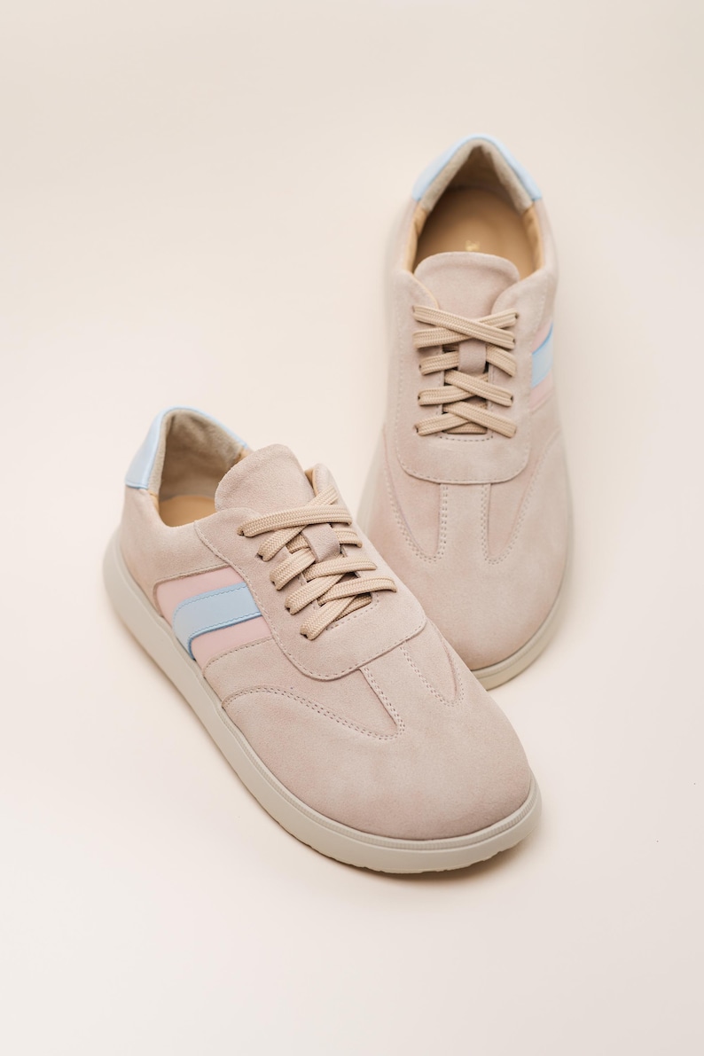 May include: A pair of light pink suede sneakers with blue and pink stripes on the sides. The laces are beige and the soles are white.